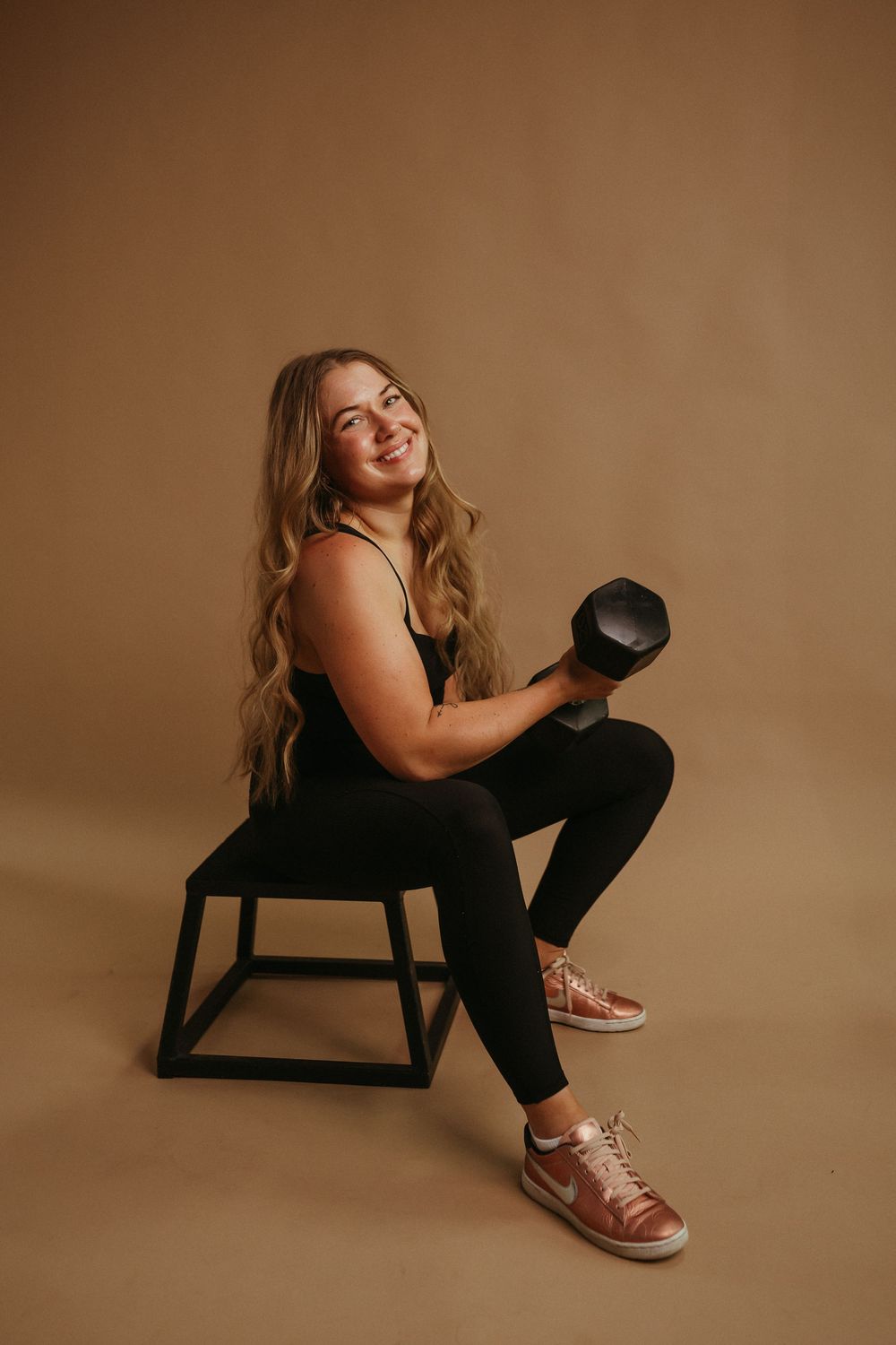Person in workout attire poses with dumbbell on wooden stool against warm brown background.