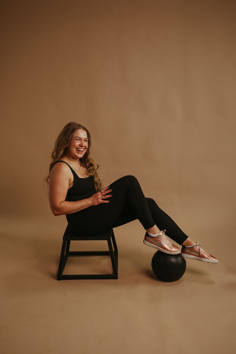 Lifestyle portrait of a person in black activewear posing on a stool with a ball against a tan studio backdrop.