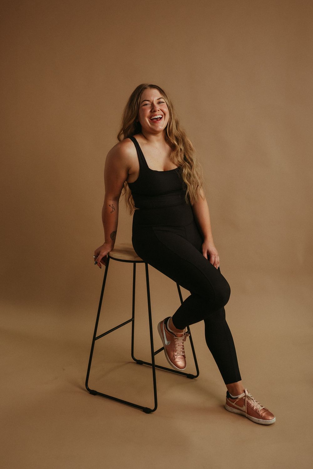 Candid studio portrait of a person wearing activewear and sneakers sitting on a metal stool on tan background.