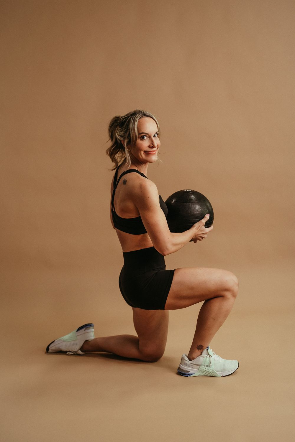 Athletic person in black workout attire performs a lunge exercise while holding a medicine ball against beige background.