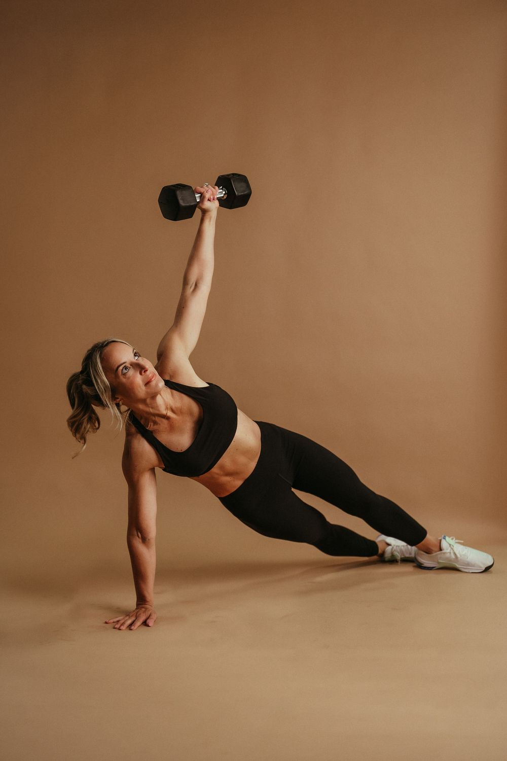 Fitness model performing side plank with dumbbell press exercise on beige background in athletic wear.