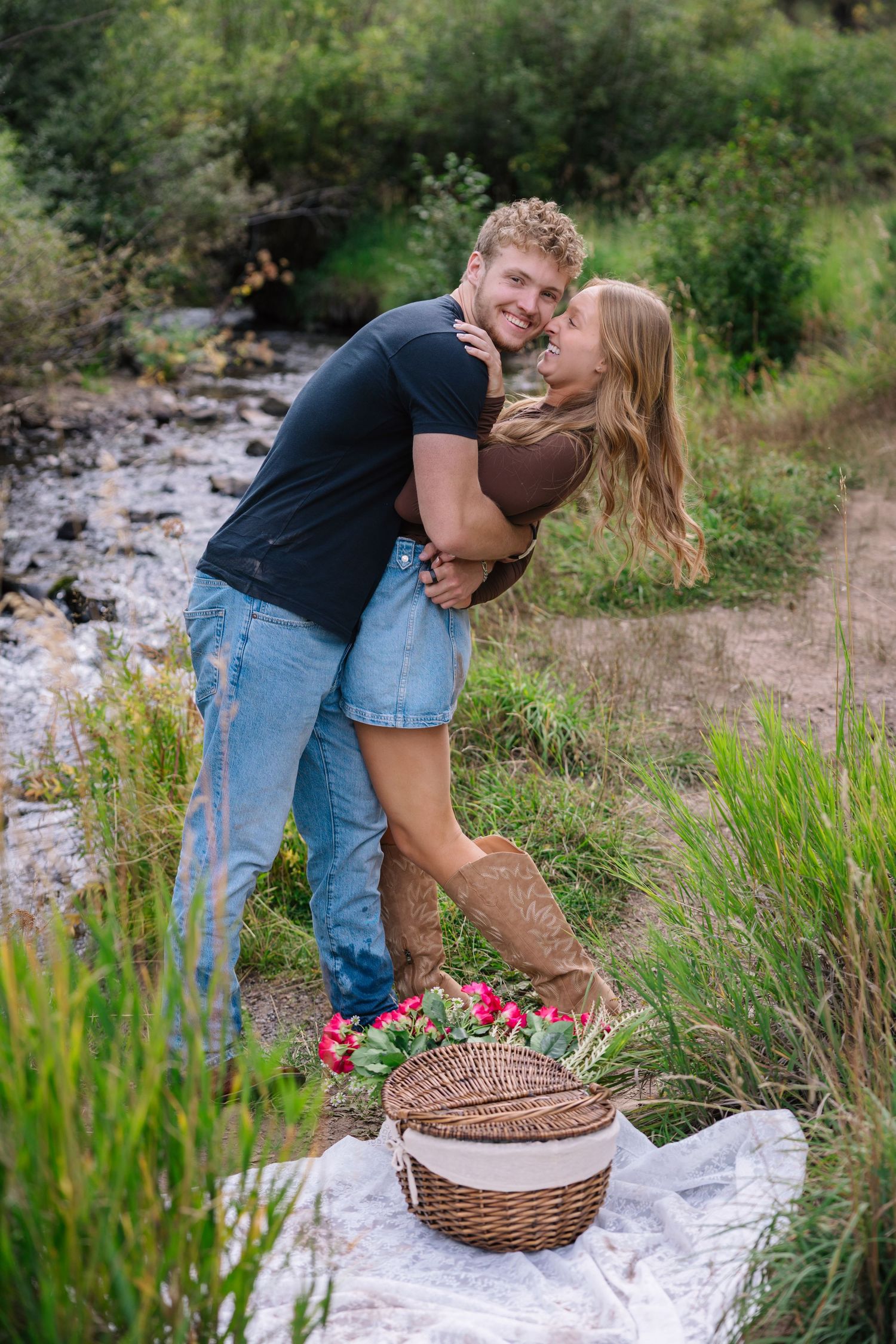 A couple embraces by a stream with a picnic basket and blanket laid out near wildflowers and grass.