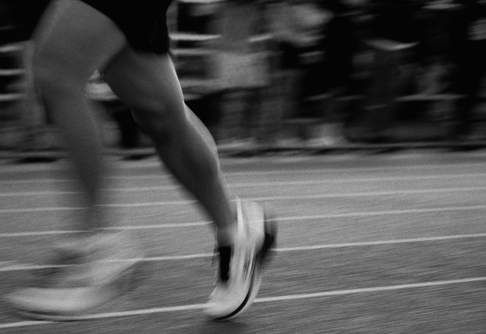 Motion-blurred black and white image showing running legs and feet on an athletic track during a race.