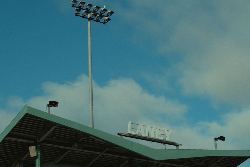 Stadium field light arrays illuminated against a cloudy twilight blue sky with stadium facilities in the background.