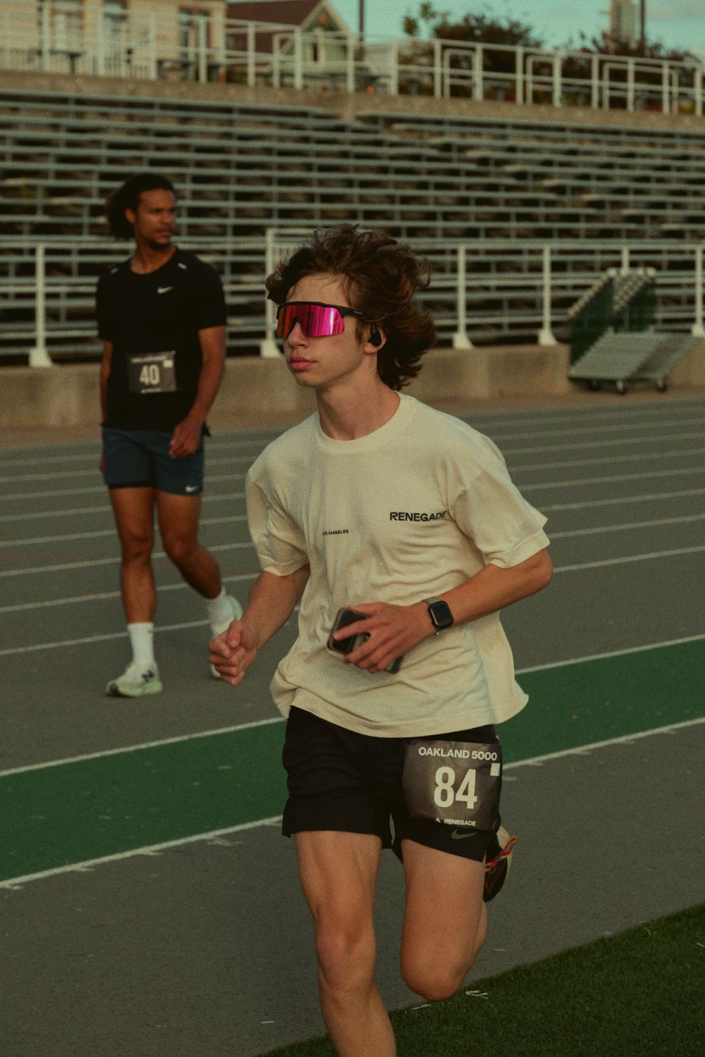 Runner with pink sunglasses and race number 84 competes on a track with stadium bleachers in background.