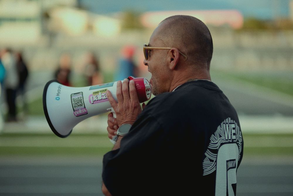 A person in sunglasses and a black shirt speaking into a white megaphone at a sports field.