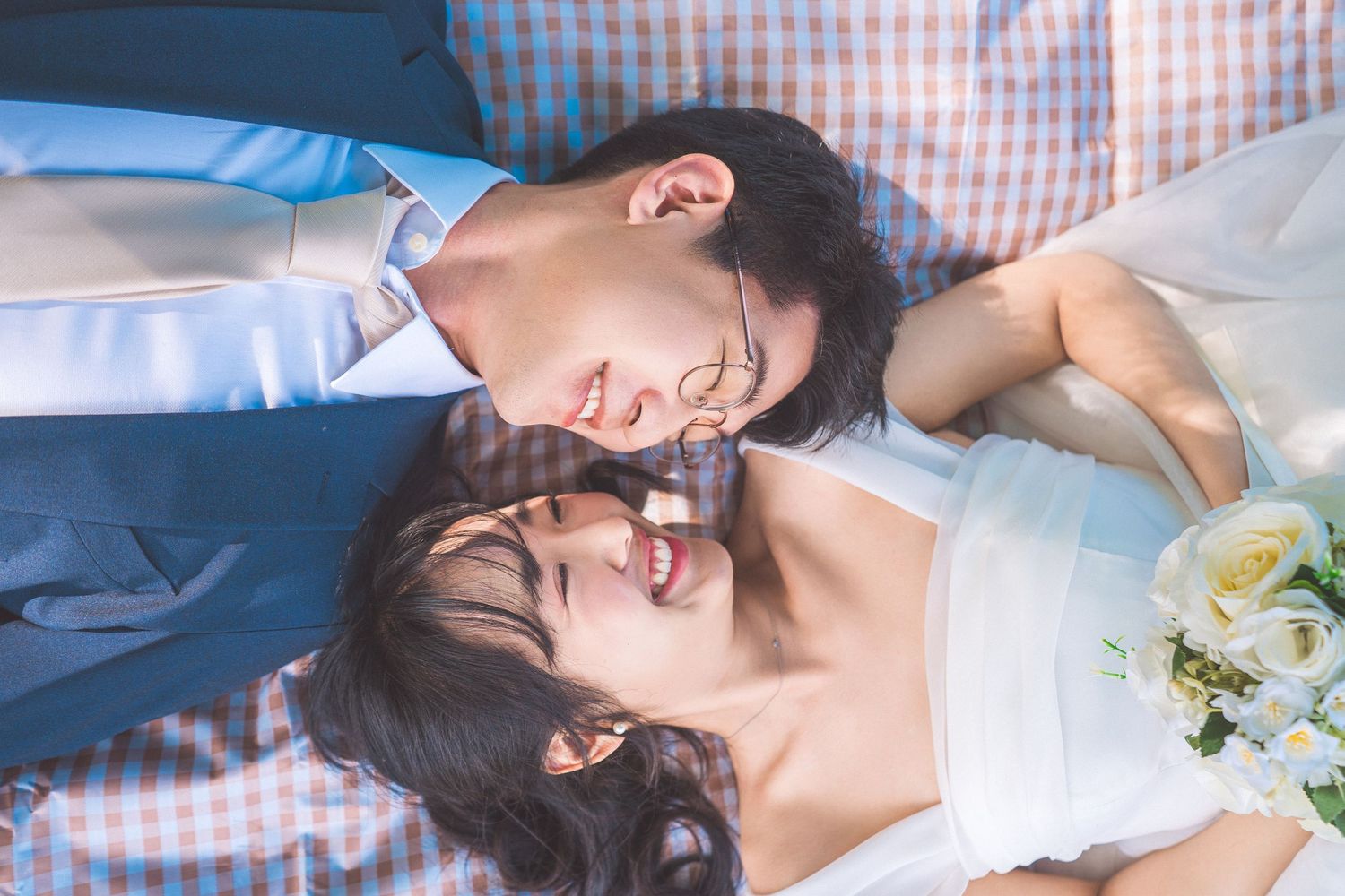 A newlywed couple lying down together with white flowers and formal wedding attire on a blue checkered background.