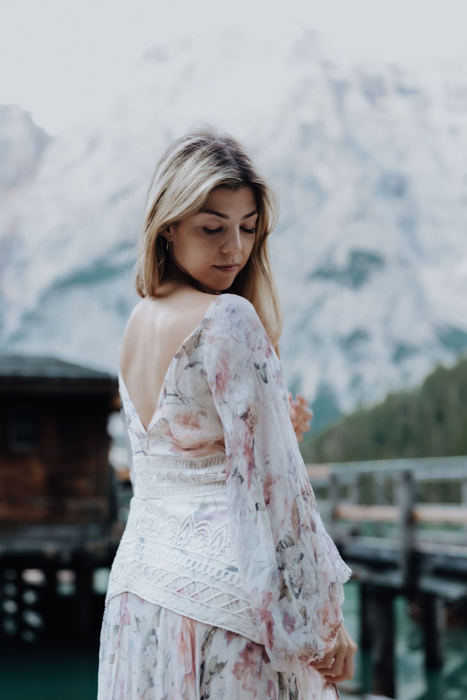 A person in a flowing white floral dress poses outdoors against a mountain backdrop.