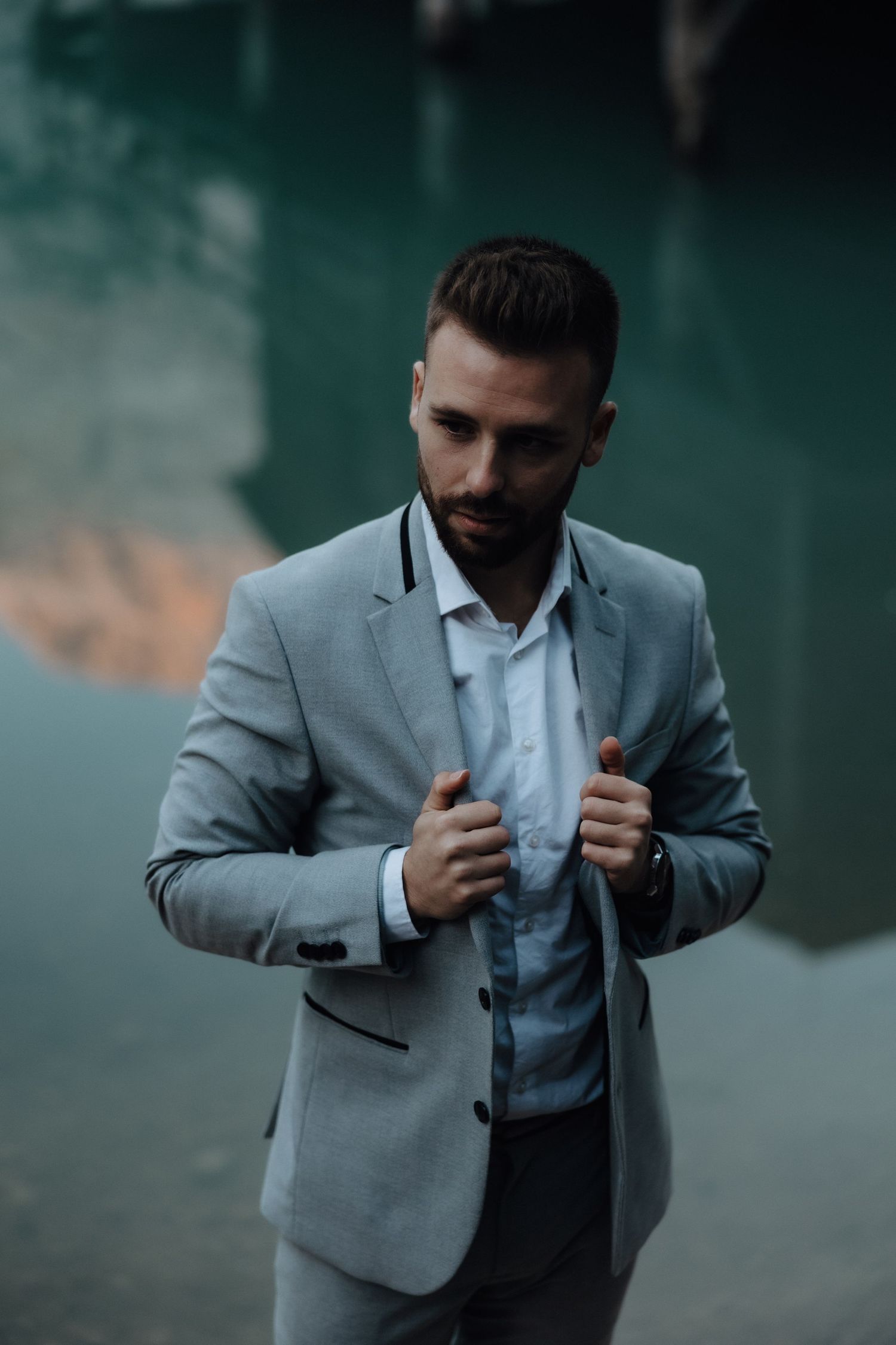 Stylish person wearing a light gray business suit adjusts his jacket against moody teal background.