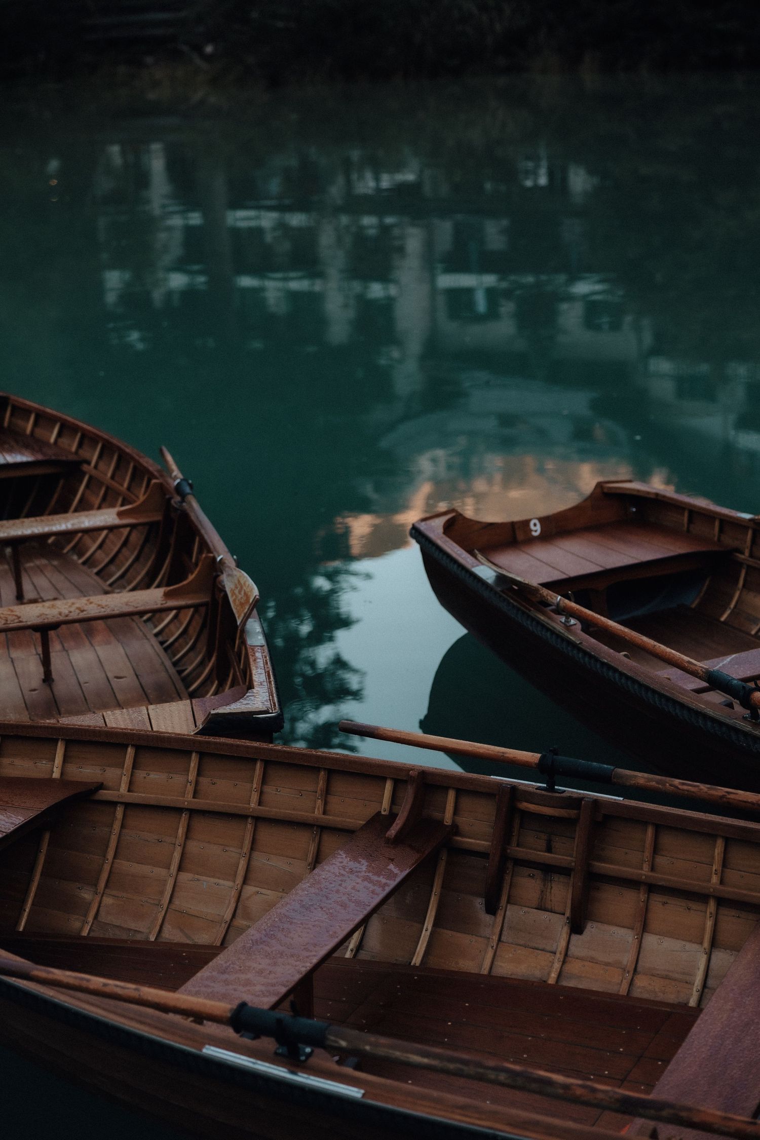 Wooden rowboats float on dark green reflective water at dusk, creating a moody and atmospheric scene.