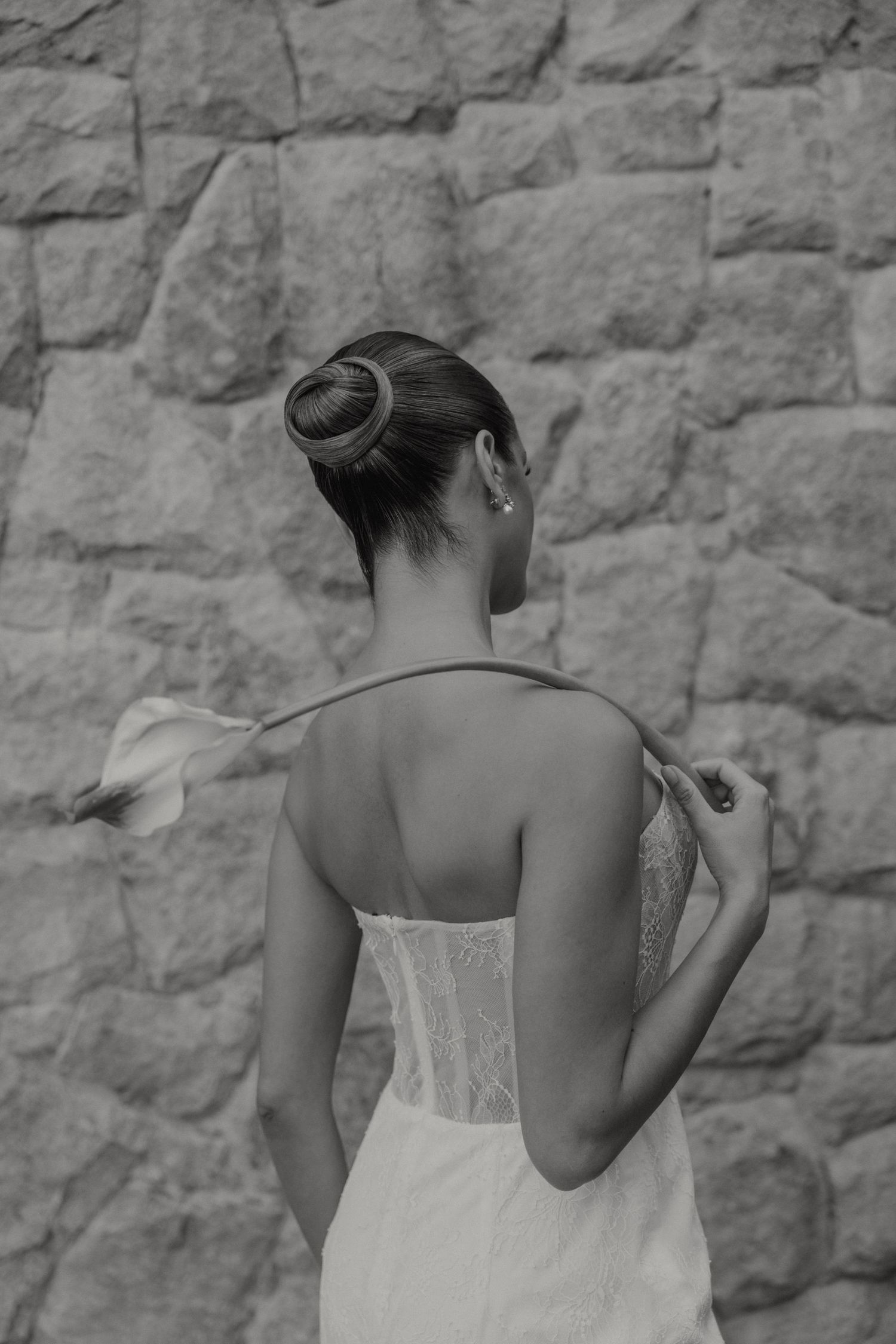 Black and white portrait of elegant bridal updo hairstyle and sheer back wedding dress against stone wall.