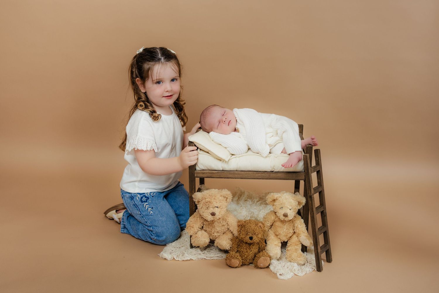 A toddler sits with a teddy bear and white blanket against a warm brown studio backdrop.