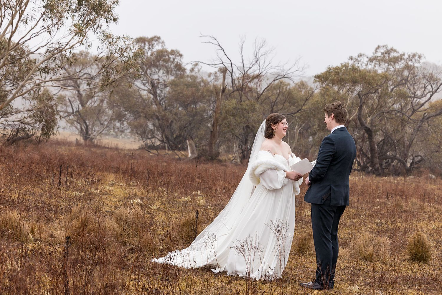 A couple in wedding attire stands together in a windswept rural field surrounded by autumn trees and foliage during their wedding vows.