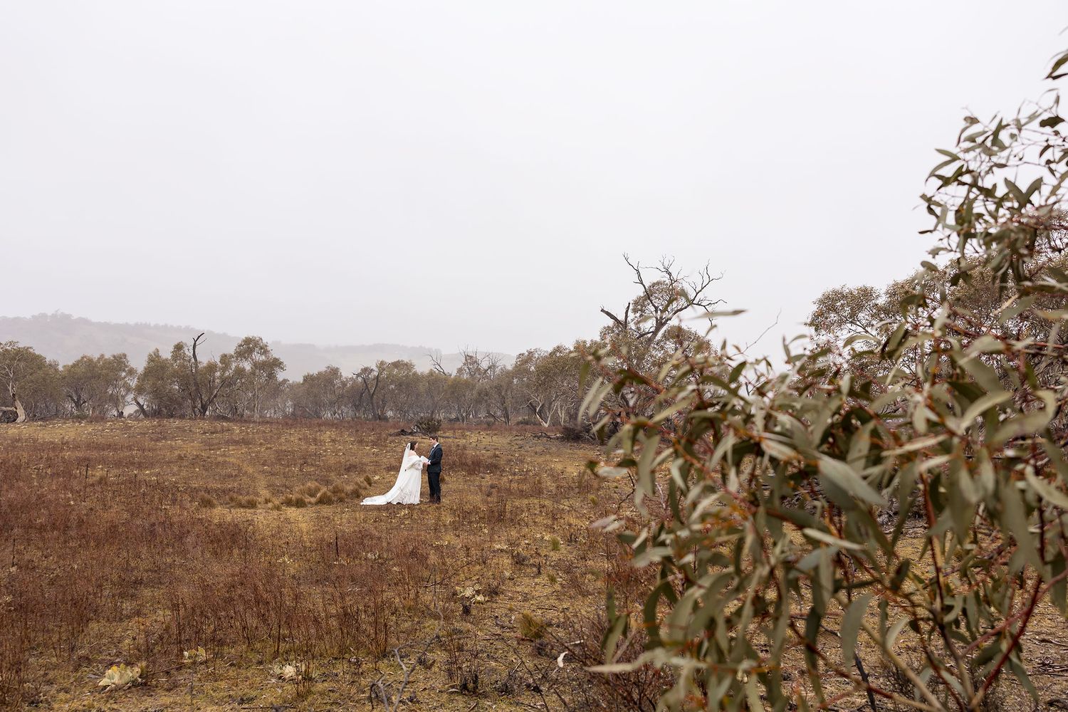 A couple in wedding attire stands together in a windswept rural field surrounded by autumn trees and foliage during their wedding vows.