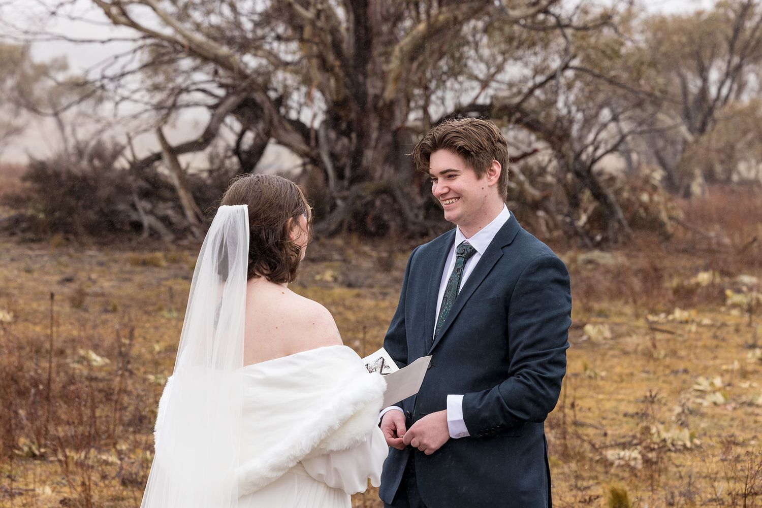 A couple in wedding attire stands together in a windswept rural field surrounded by autumn trees and foliage during their wedding vows.