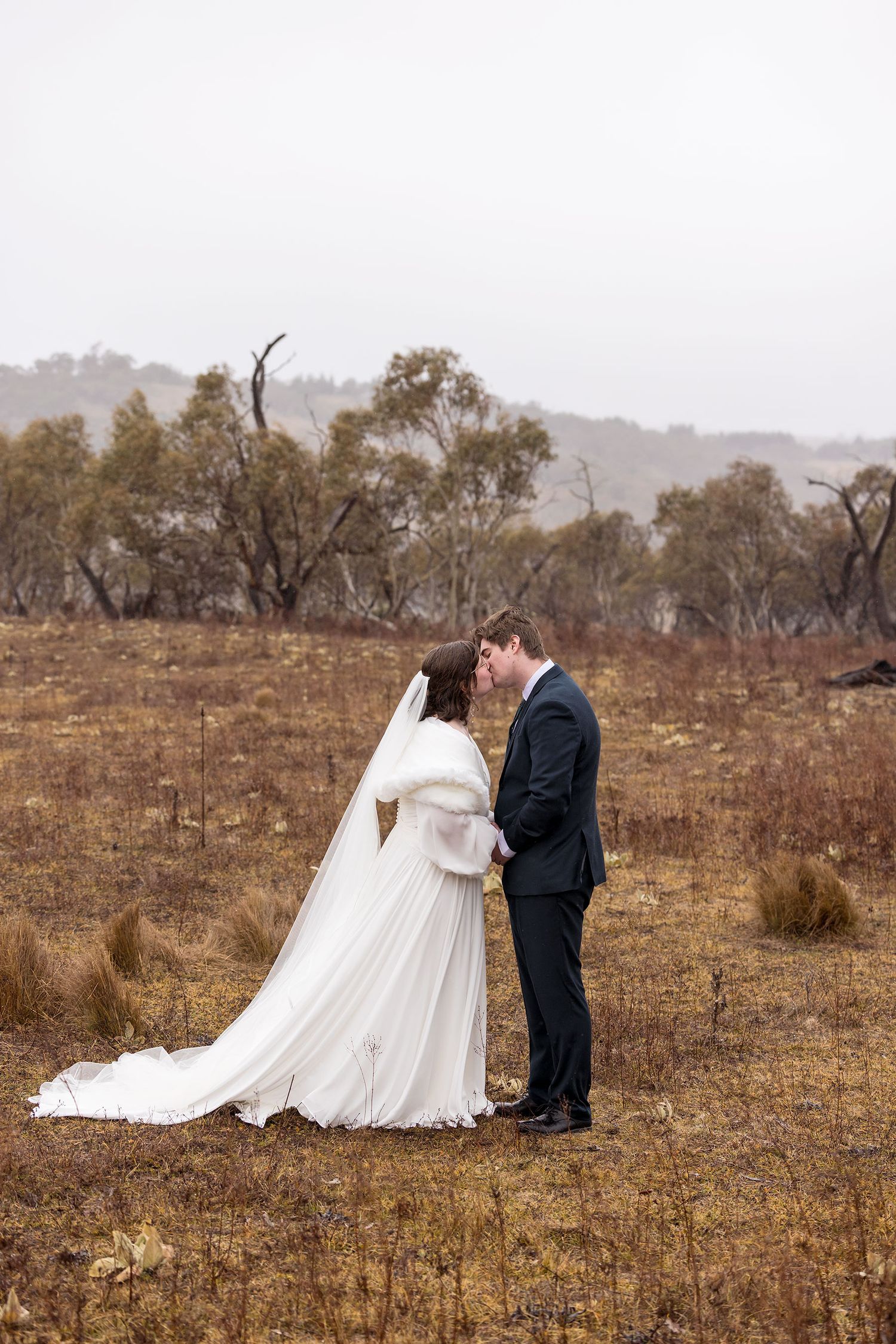 A couple in wedding attire embrace while standing in a rustic brown field with scattered trees in the background.