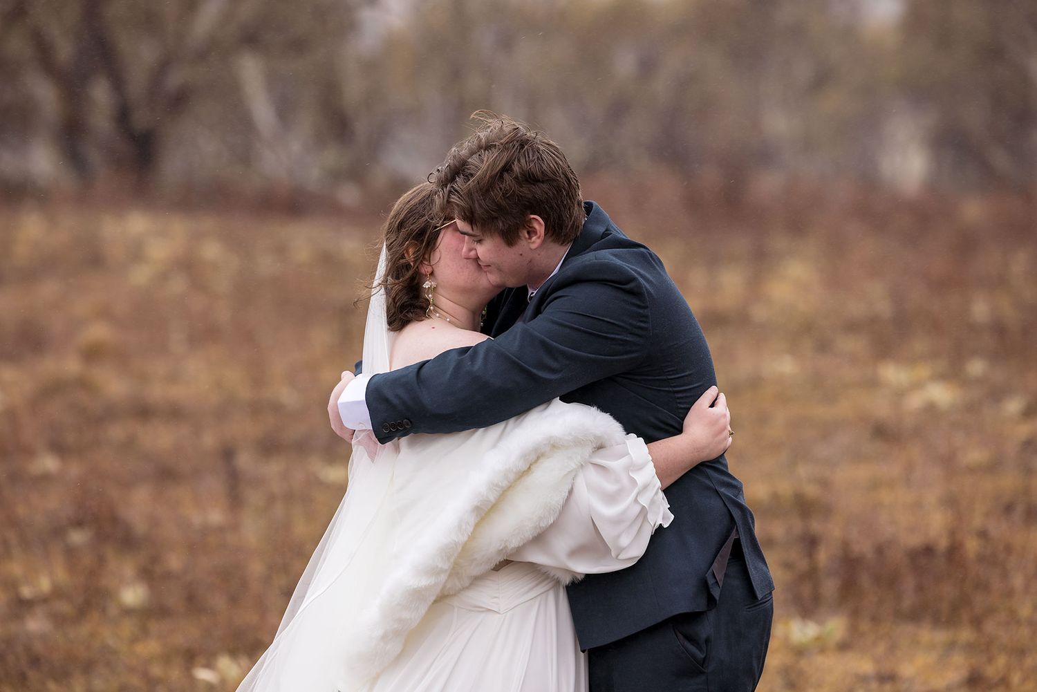 A couple shares a romantic kiss and embrace during an outdoor wedding ceremony in a late winter field in Jindabyne.