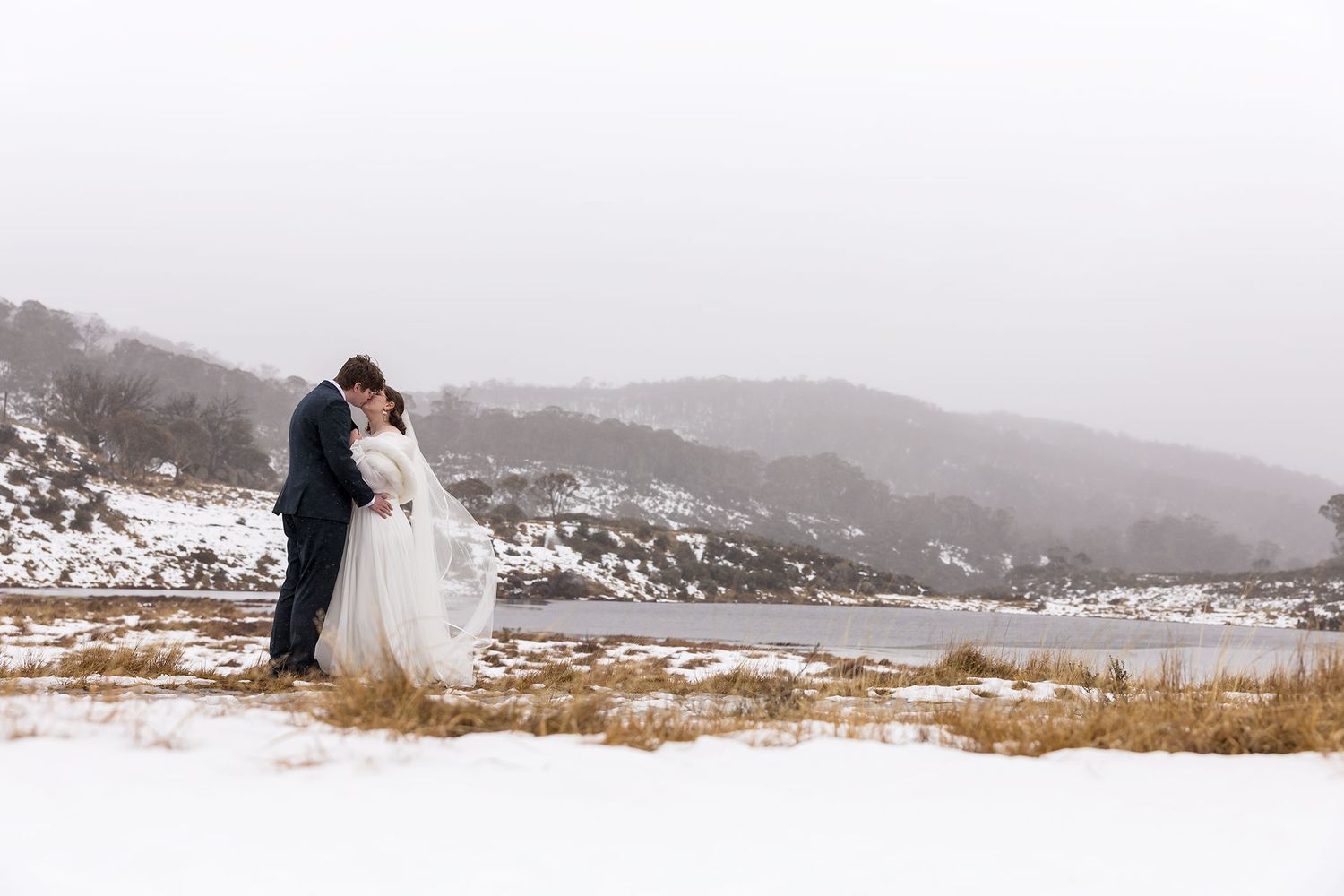 A couple in wedding attire embraces on a snowy mountain overlook on their winter wedding day.