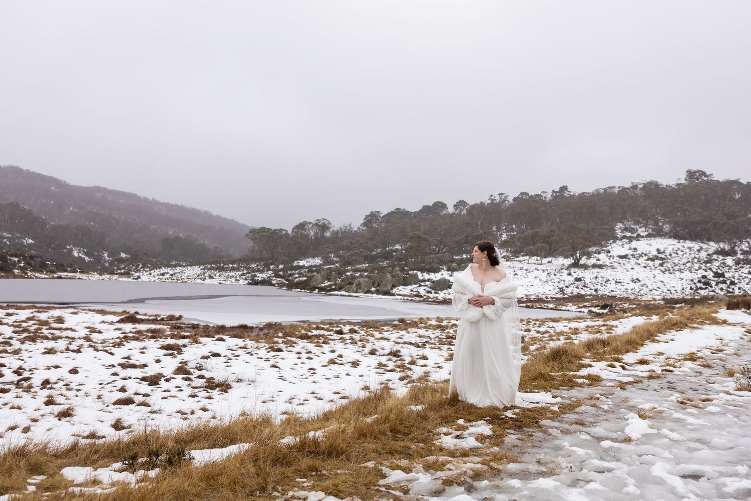 A Bride in a white 'All Who Wander' wedding dress stands in a snowy landscape overlooking a frozen lake and hills in the winter.