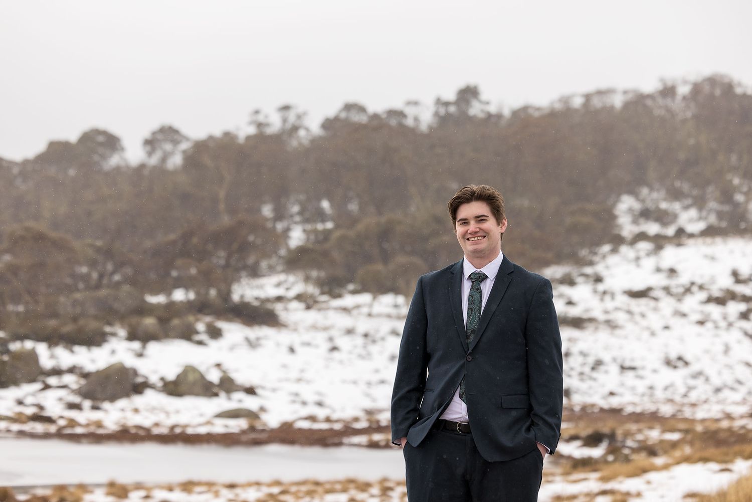 A Groom in a dark wedding suit stands on snowy terrain with trees visible on a hillside in the background at Perisher.