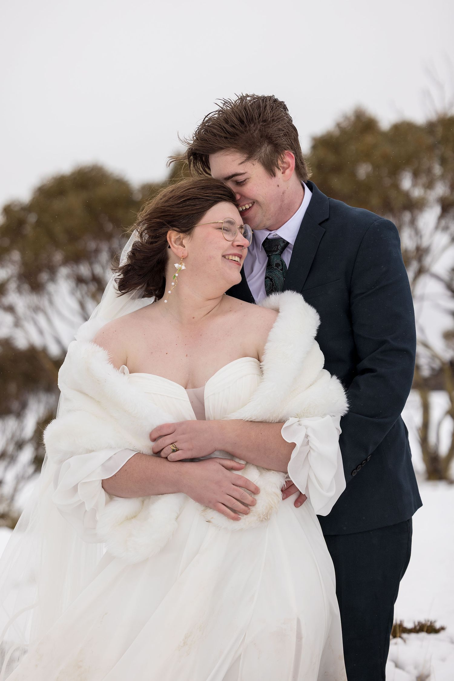 A romantic winter wedding scene with a couple embracing outdoors in the snow, the bride wearing a white gown with fur wrap.