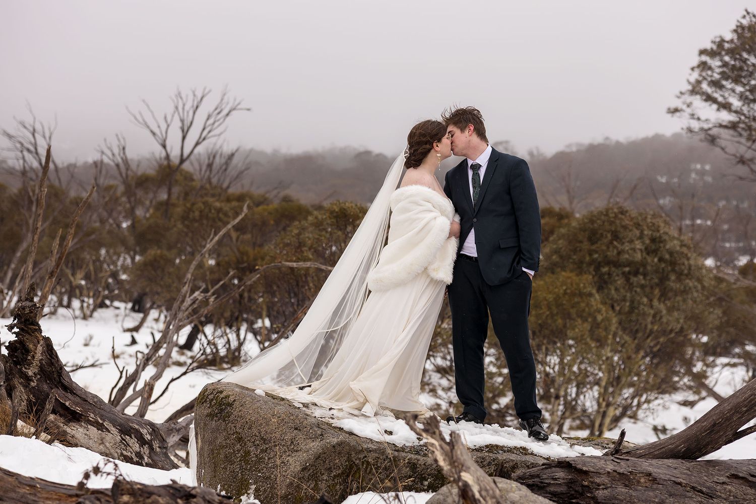 A romantic winter wedding scene with a Bride and Groom embracing outdoors in the snow, the bride wearing a white gown with fur wrap.