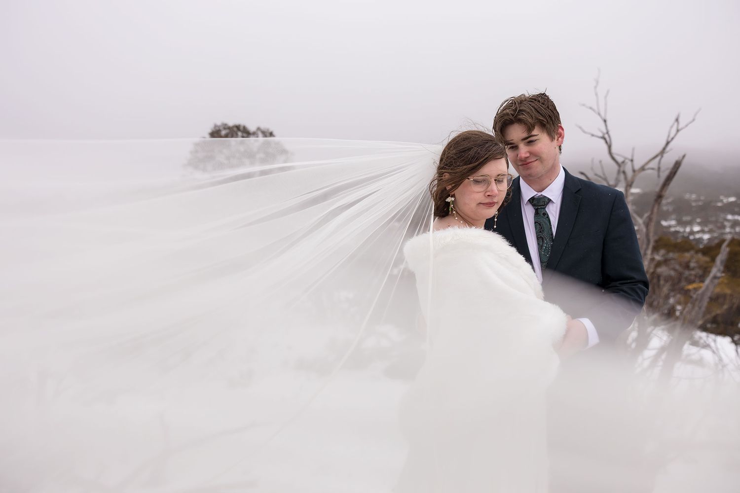 A couple in wedding attire stands close together in a misty, ethereal outdoor setting as the bride's veil billows dramatically in the snow.