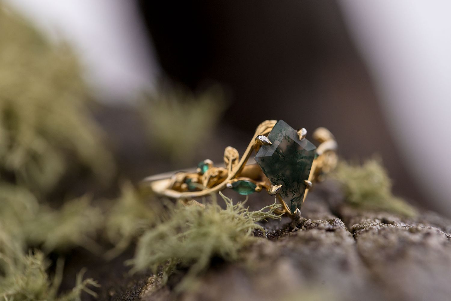Elegant gold ring with dark green gemstone photographed in macro against blurred mossy background.