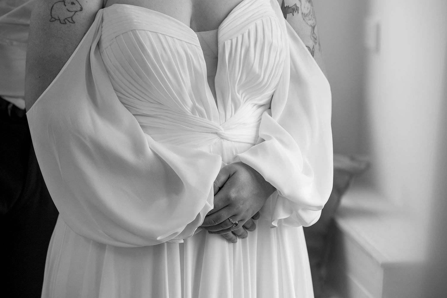 Black and white photo showing a Bride in an off-shoulder white 'All Who Wander' gown with a detailed tattoo visible on their upper arm.