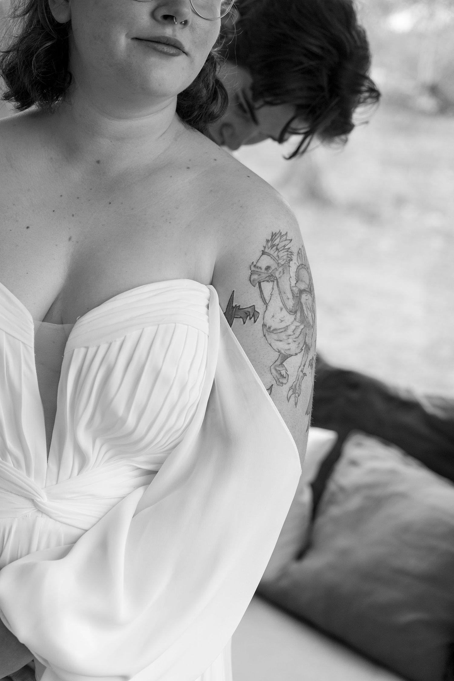 Black and white photo showing a Bride in an off-shoulder white 'All Who Wander' gown with a detailed tattoo visible on their upper arm.