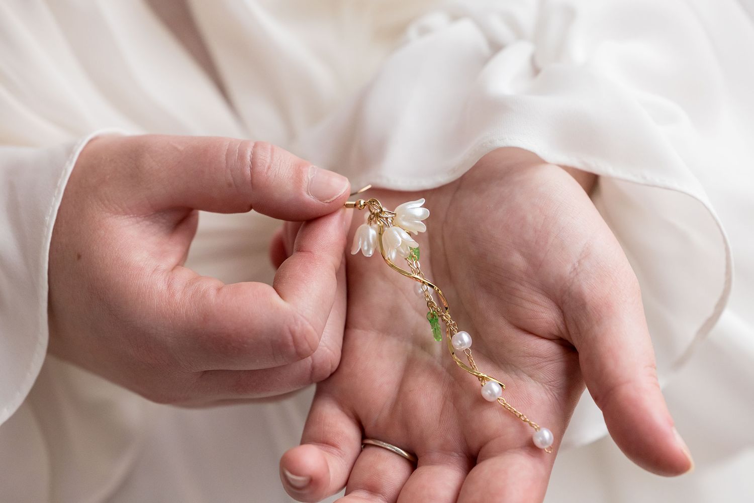 Hands holding delicate pearl and crystal jewelry against a soft white fabric background.