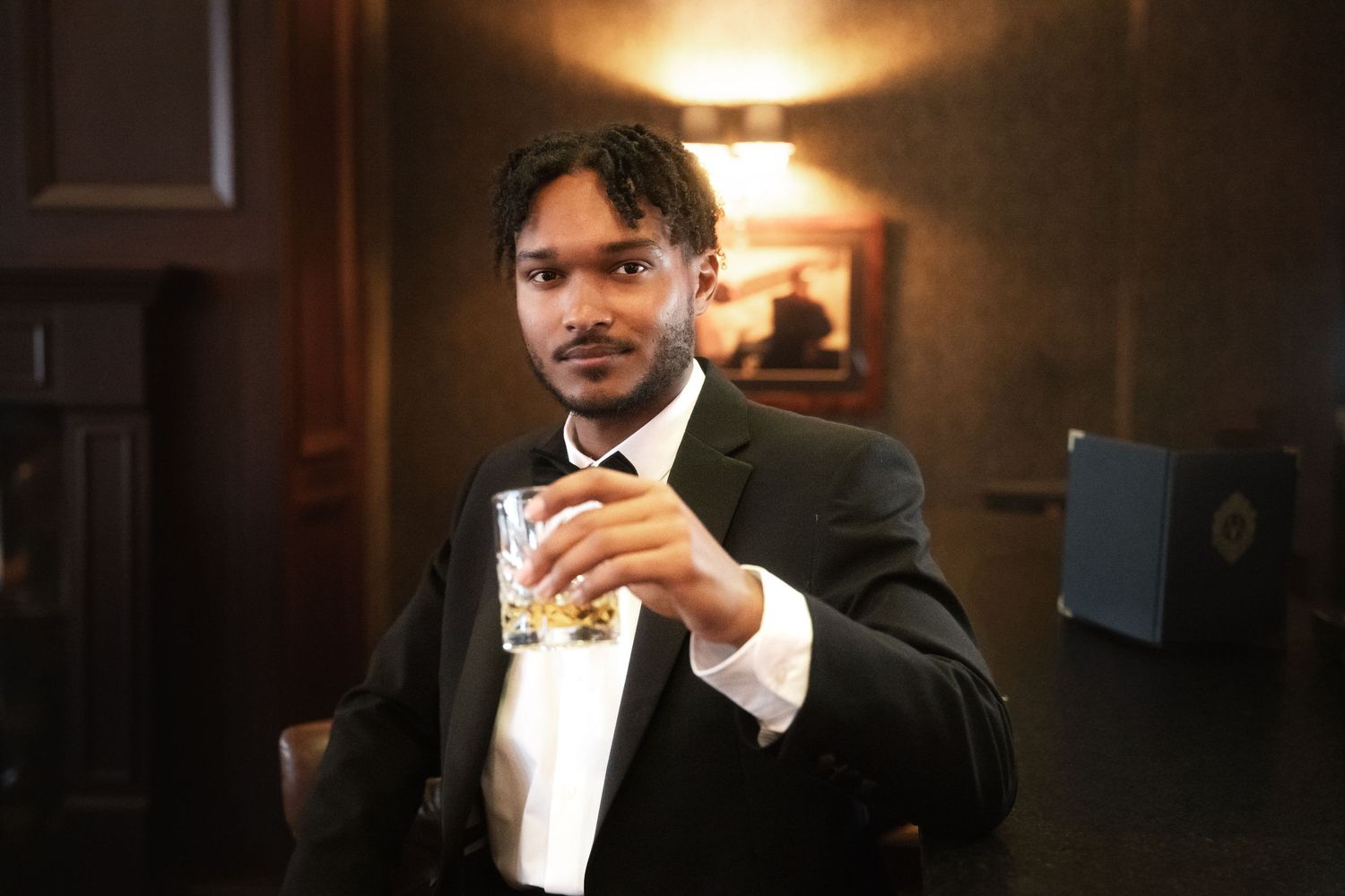 A person in a tuxedo with a bowtie adjusts their collar in a dimly lit room.