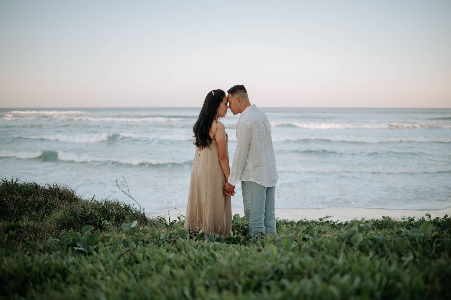A couple shares a romantic moment on a grassy cliff overlooking the ocean waves at sunset.