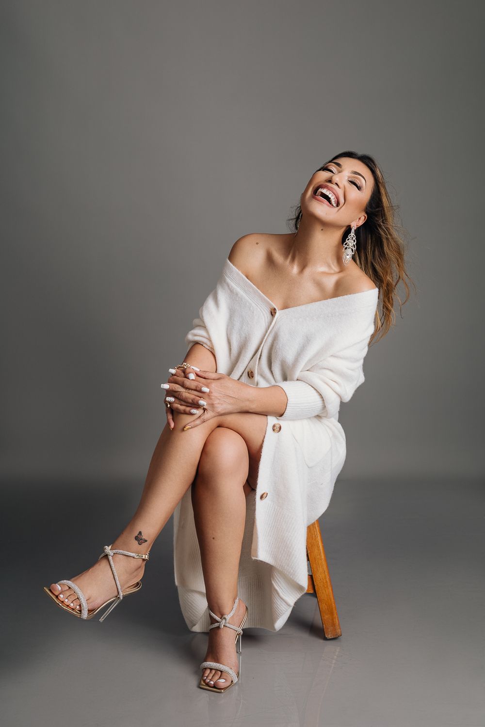 Fashion portrait of model in white sweater dress sitting on wooden chair with expressive laughter.