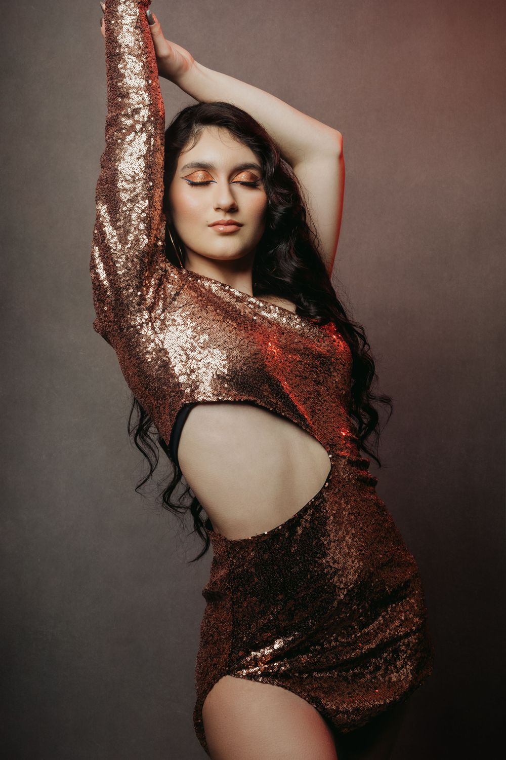 Model in copper sequin dress strikes graceful pose with arm raised.