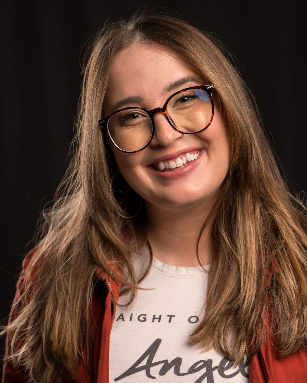 Young person with long brown hair and glasses smiling in casual attire against dark background.