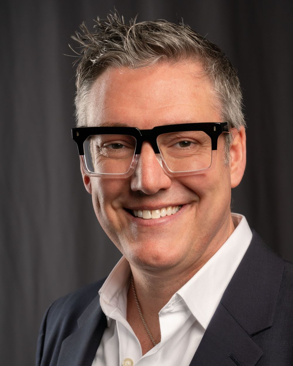 Professional headshot of person wearing thick-framed glasses and suit jacket smiling warmly against dark background.