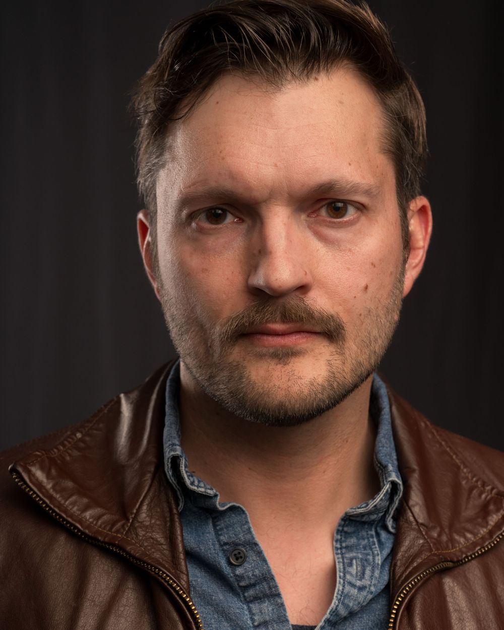 Portrait of person in leather jacket and denim shirt with facial hair against dark backdrop.