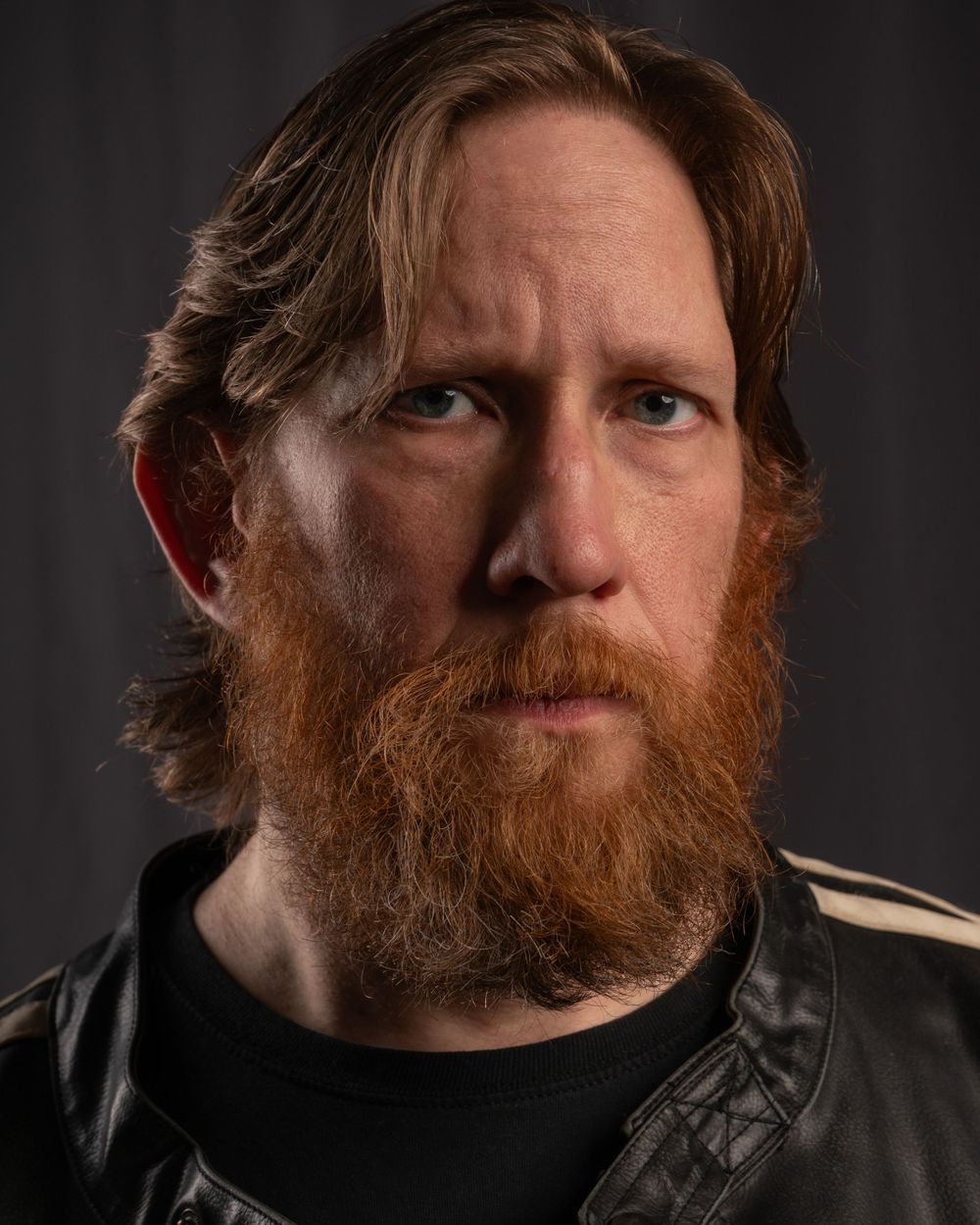 Dramatic portrait of person with red beard wearing black leather jacket against dark backdrop.