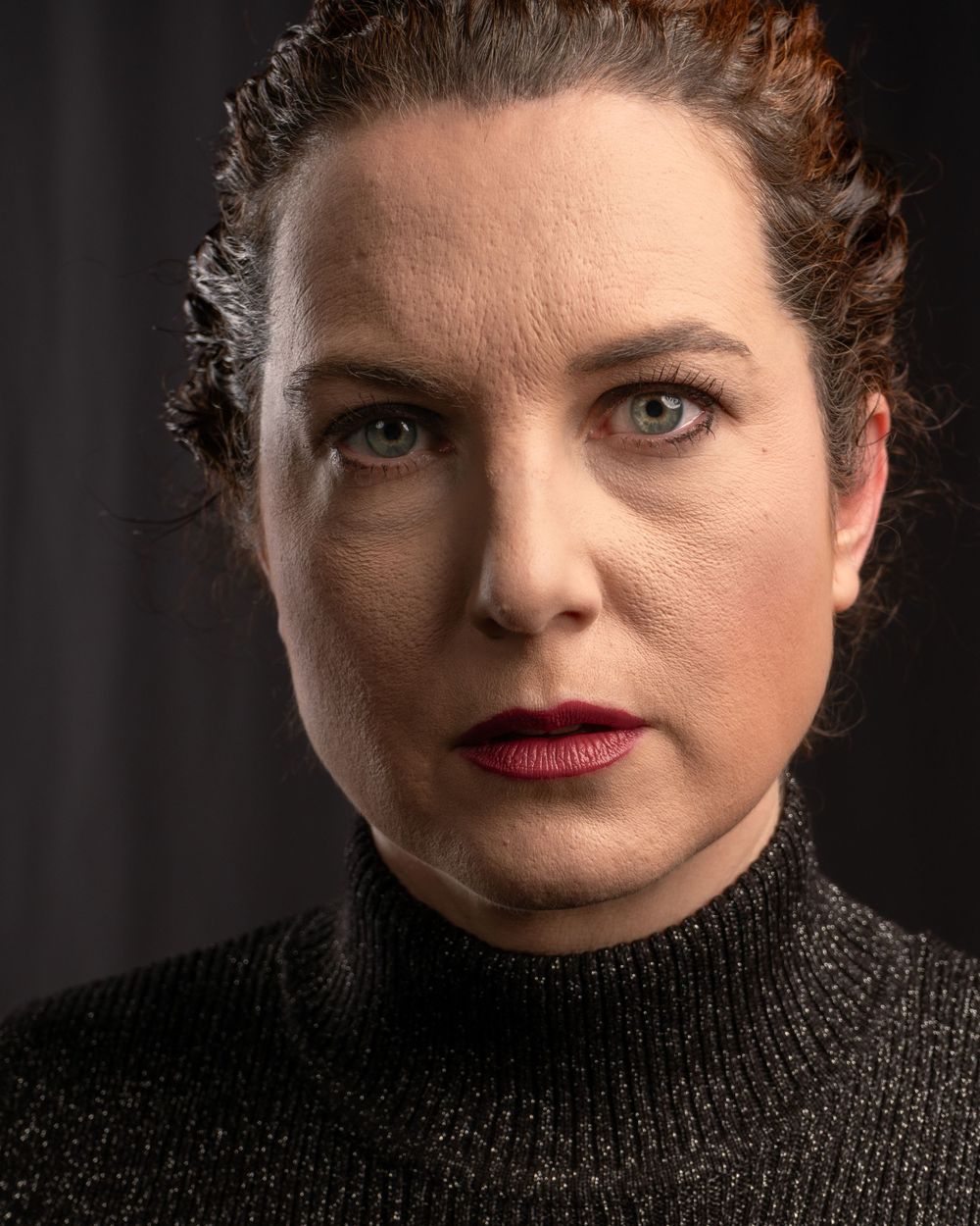 Close-up portrait of person in black turtleneck with dark lipstick against dark background.