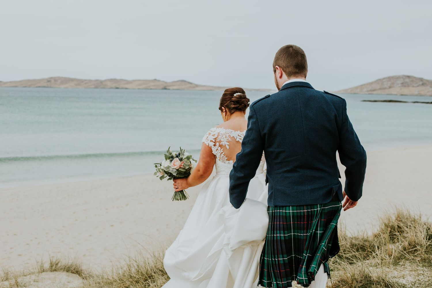 Married couple in traditional Scottish wedding attire walk along a scenic coastal landscape during their ceremony.