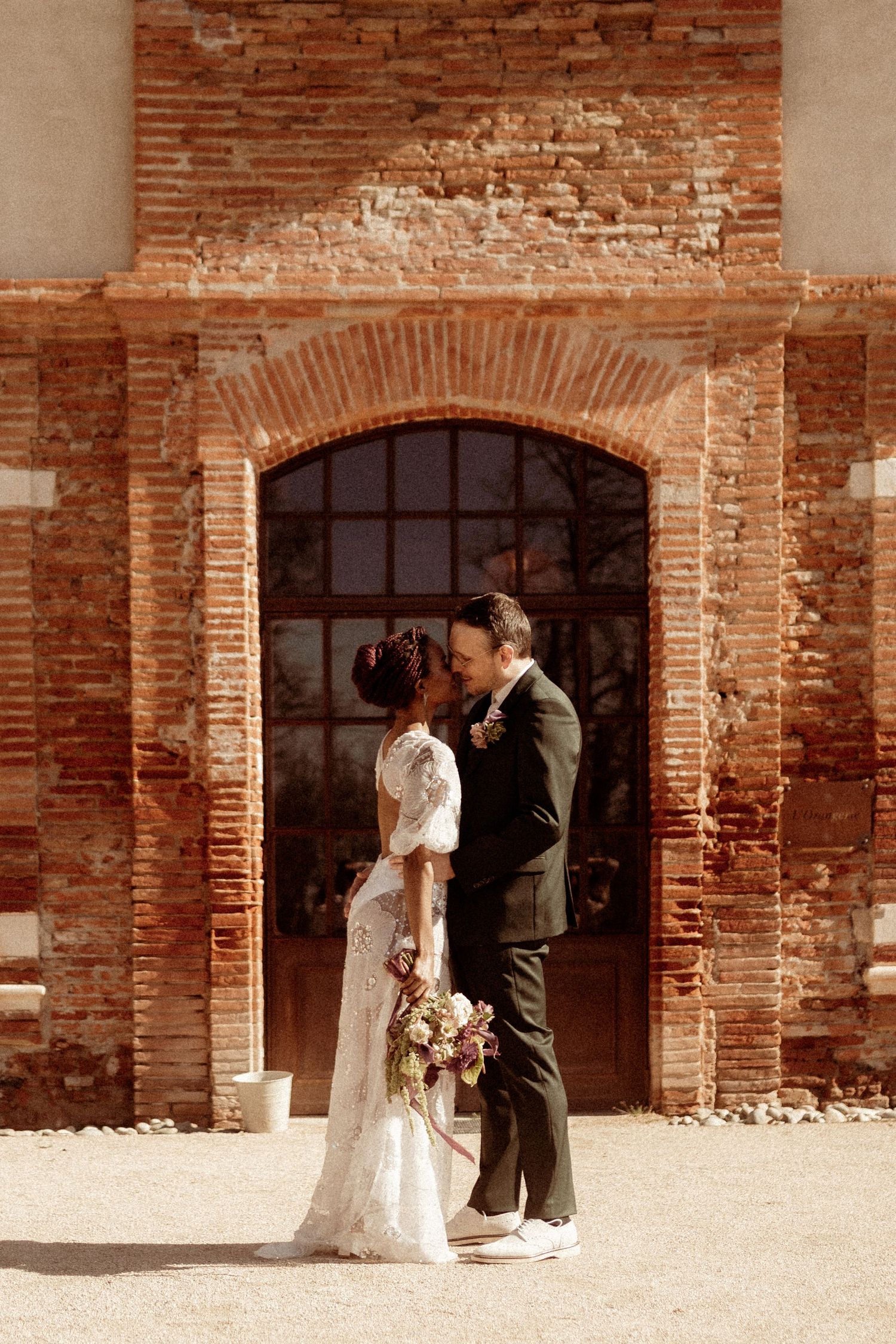 A couple in wedding attire embraces in front of a rustic brick archway at a historic venue.