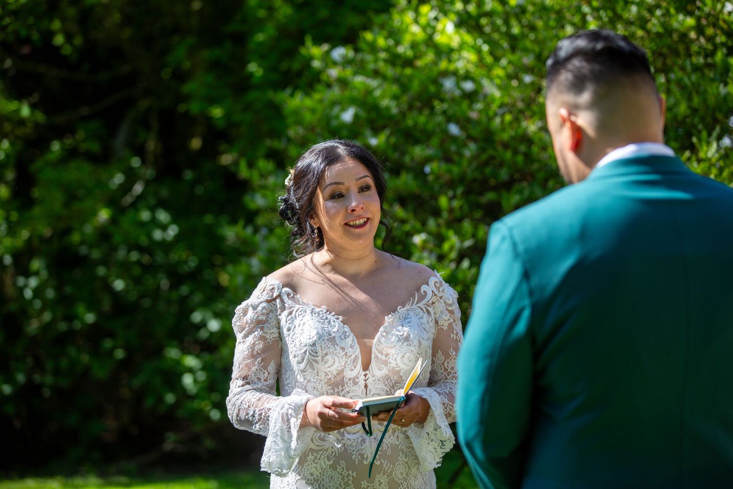 Outdoor wedding ceremony shows couple exchanging vows in a garden setting with green foliage in background.