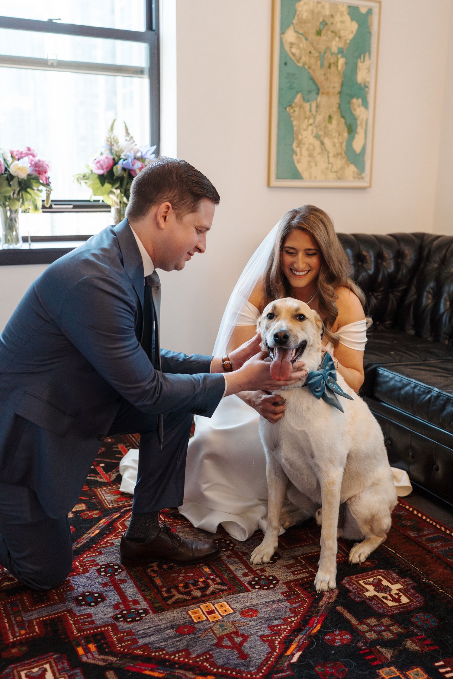 A couple in formal attire pets their dog wearing a blue bowtie while sitting on a leather couch.