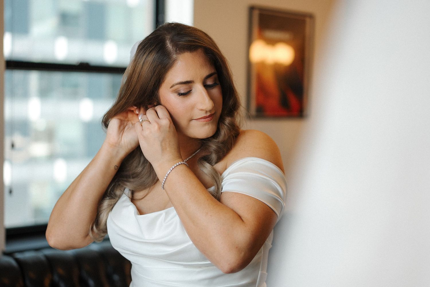 Woman in an elegant white dress poses thoughtfully near large windows in a romantic indoor setting.
