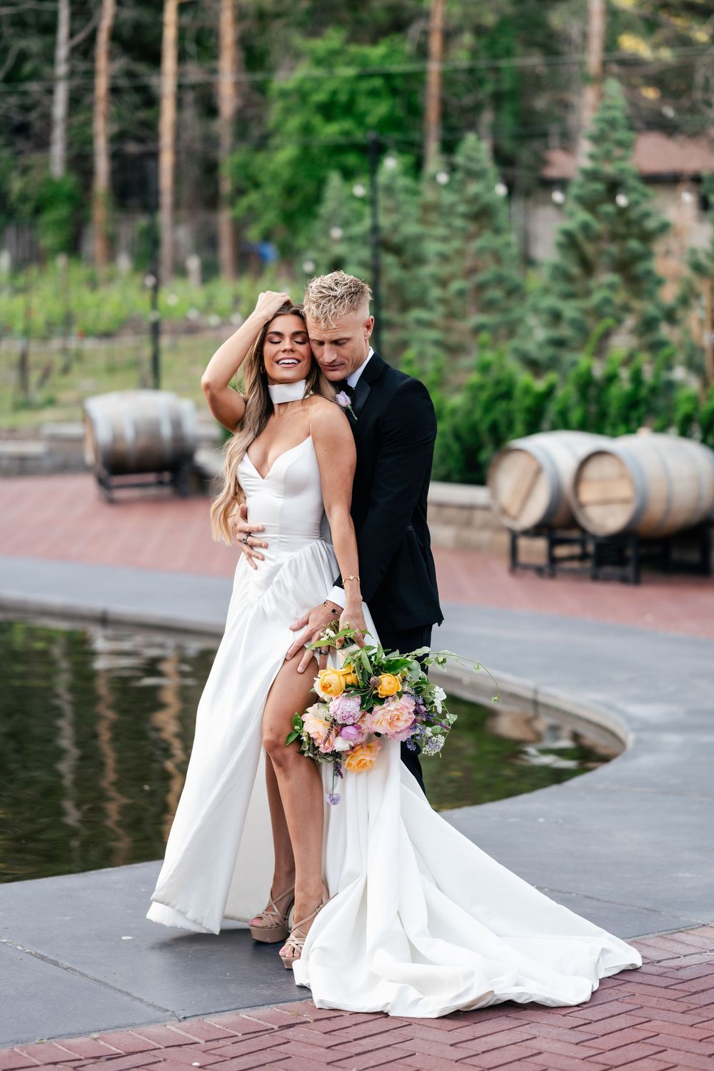 A couple in wedding attire share a romantic moment by wine barrels and a pond with pine trees in the background.