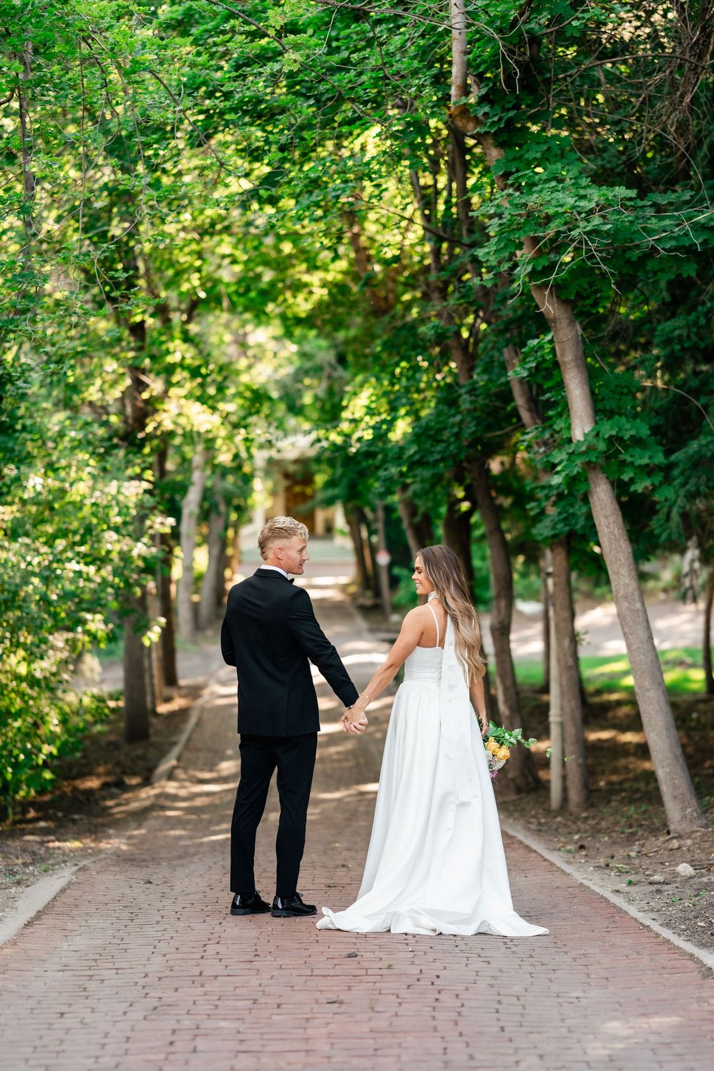 Couple walking down tree-lined brick path in wedding attire against forest backdrop.