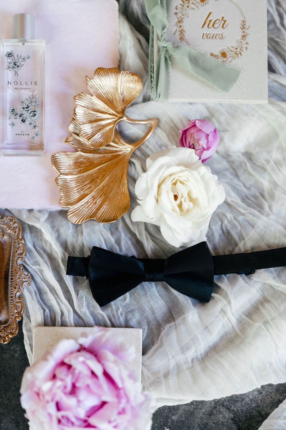 Wedding details including a perfume bottle, gold leaf decor, black bowtie, and delicate roses on light fabric.