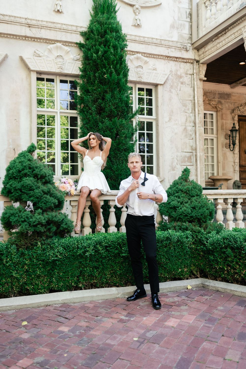 Elegant couple posing playfully on a brick patio in front of a stone mansion with landscaped cypress trees.