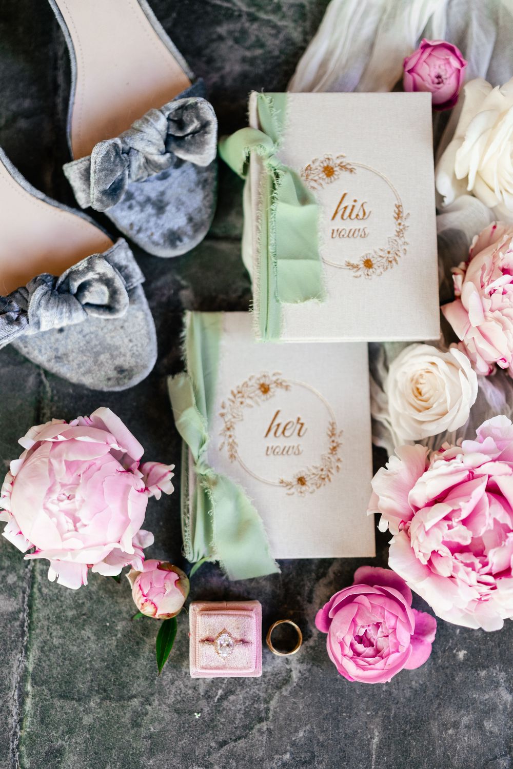 Wedding details flatlay with His and Hers vow books, pink roses, velvet ring box and grey shoes on dark background.